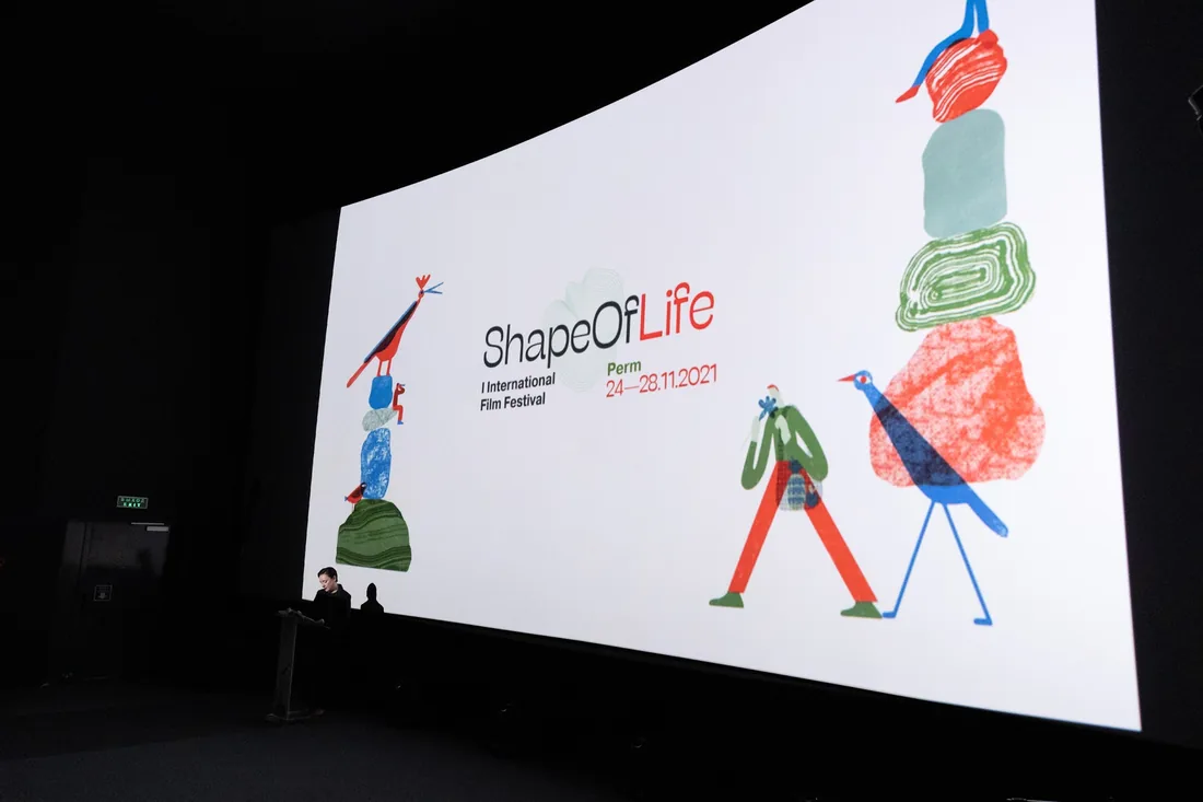 A cinema screen filling a dark auditorium with the English ShapeOfLife lockup and the stacked-stone illustration on either side, a presenter at a lectern below.