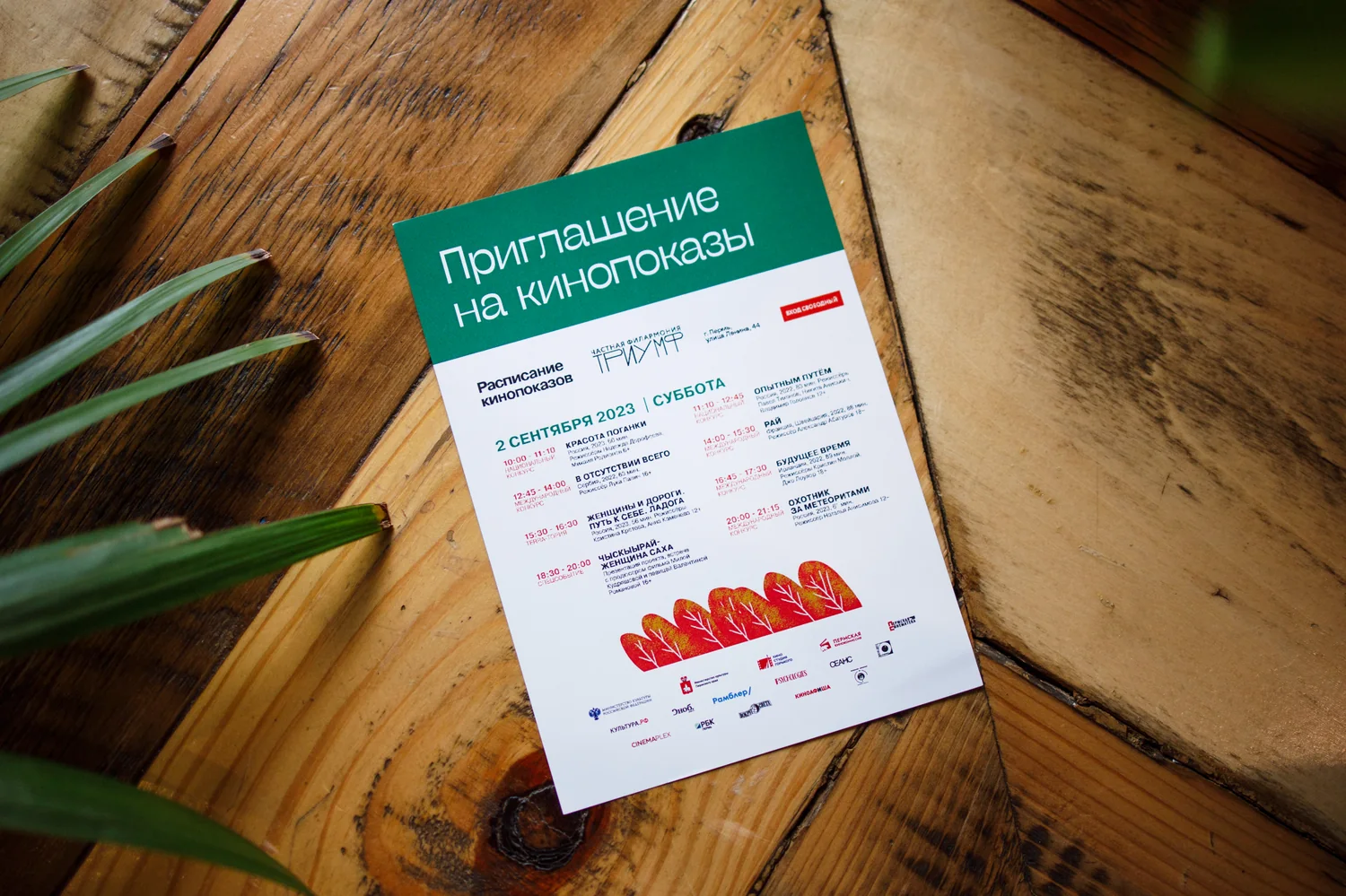The printed screening invitation, mint green with a red schedule block, photographed on a wooden table.