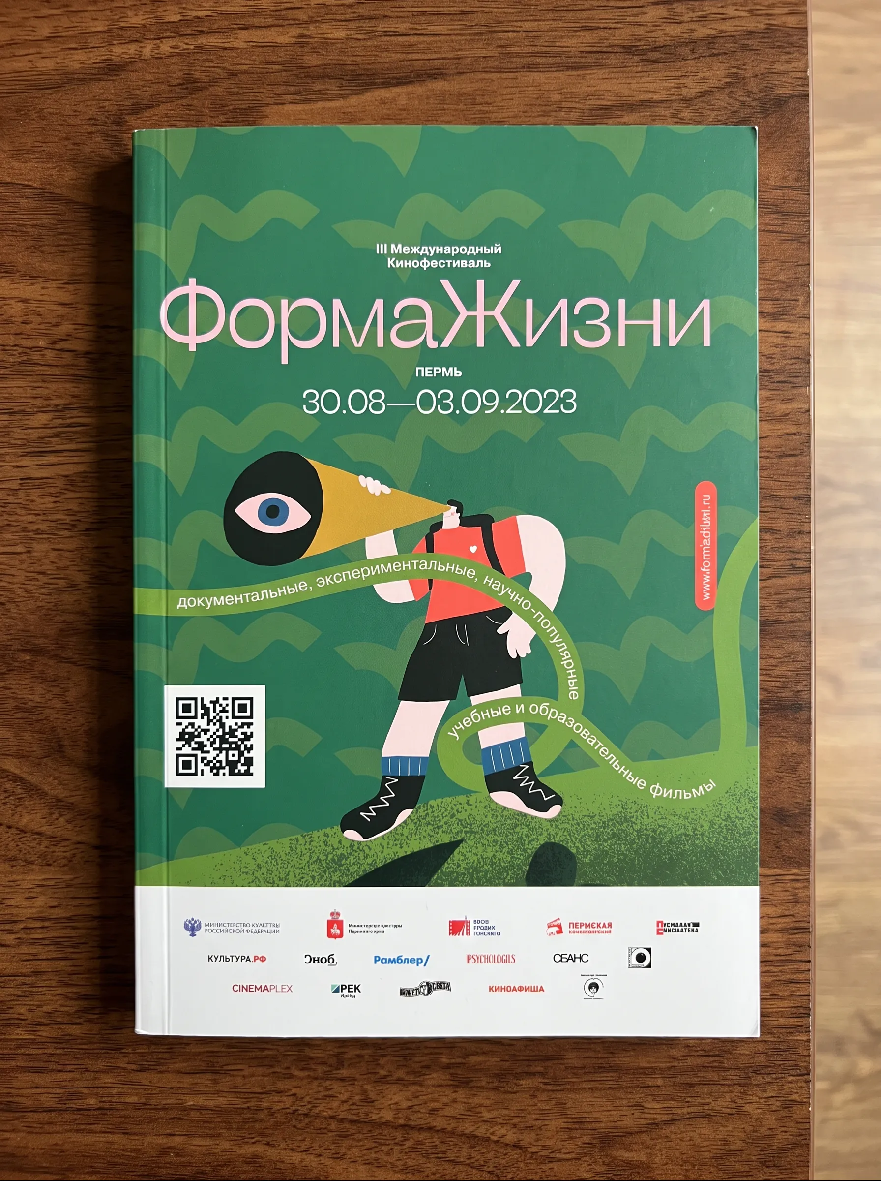 The printed festival catalogue on a wooden table, green cover with the figure and megaphone illustration and a ribbon carrying the programme categories.