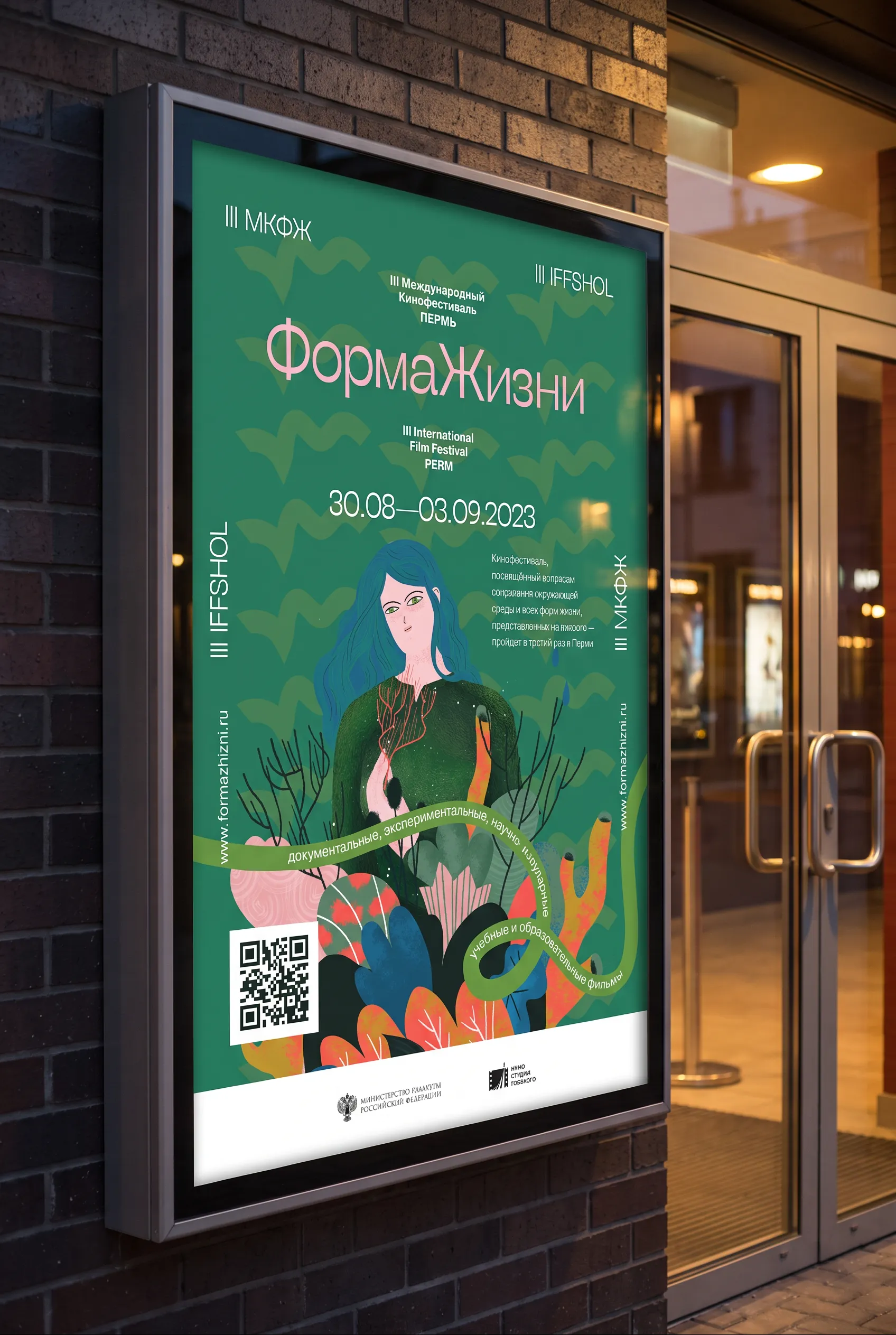 The 2023 key poster in a lit street case on a brick wall at dusk: a woman with blue hair painted into a green landscape, the wordmark above and a ribbon carrying the dates.