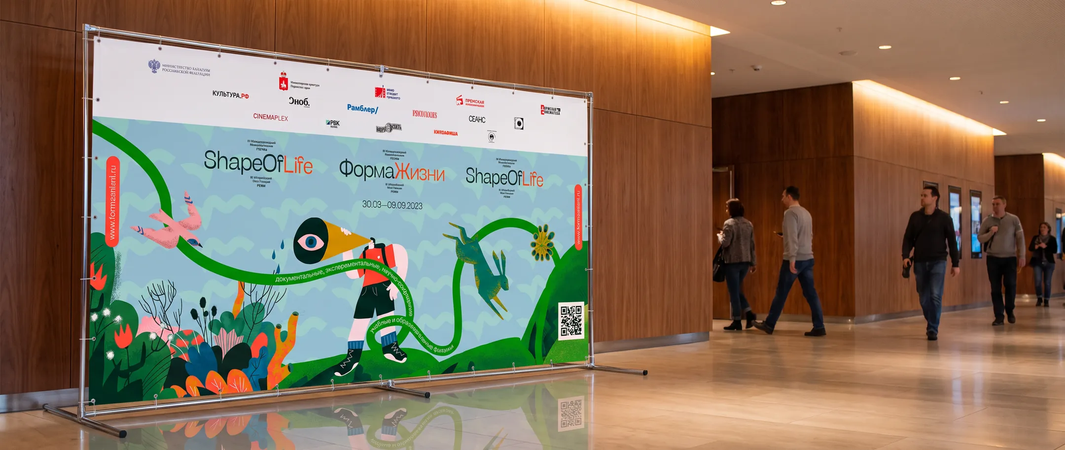 The festival press wall on a banner stand in a wood-panelled foyer, visitors walking past, printed with the illustration and both language lockups.