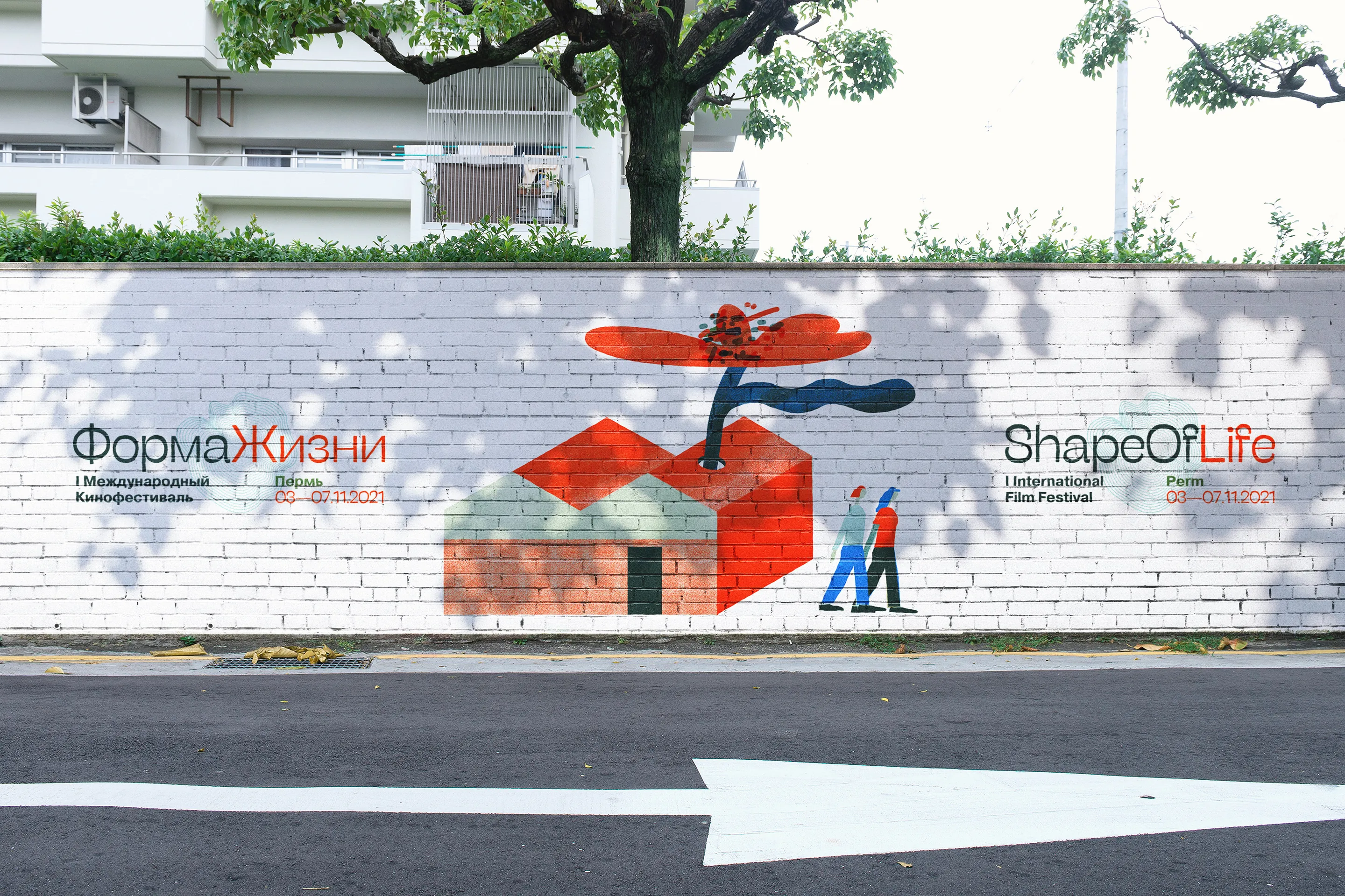 A painted street mural for the Shape of Life film festival: a red house with a tree growing out of it and two small figures walking.