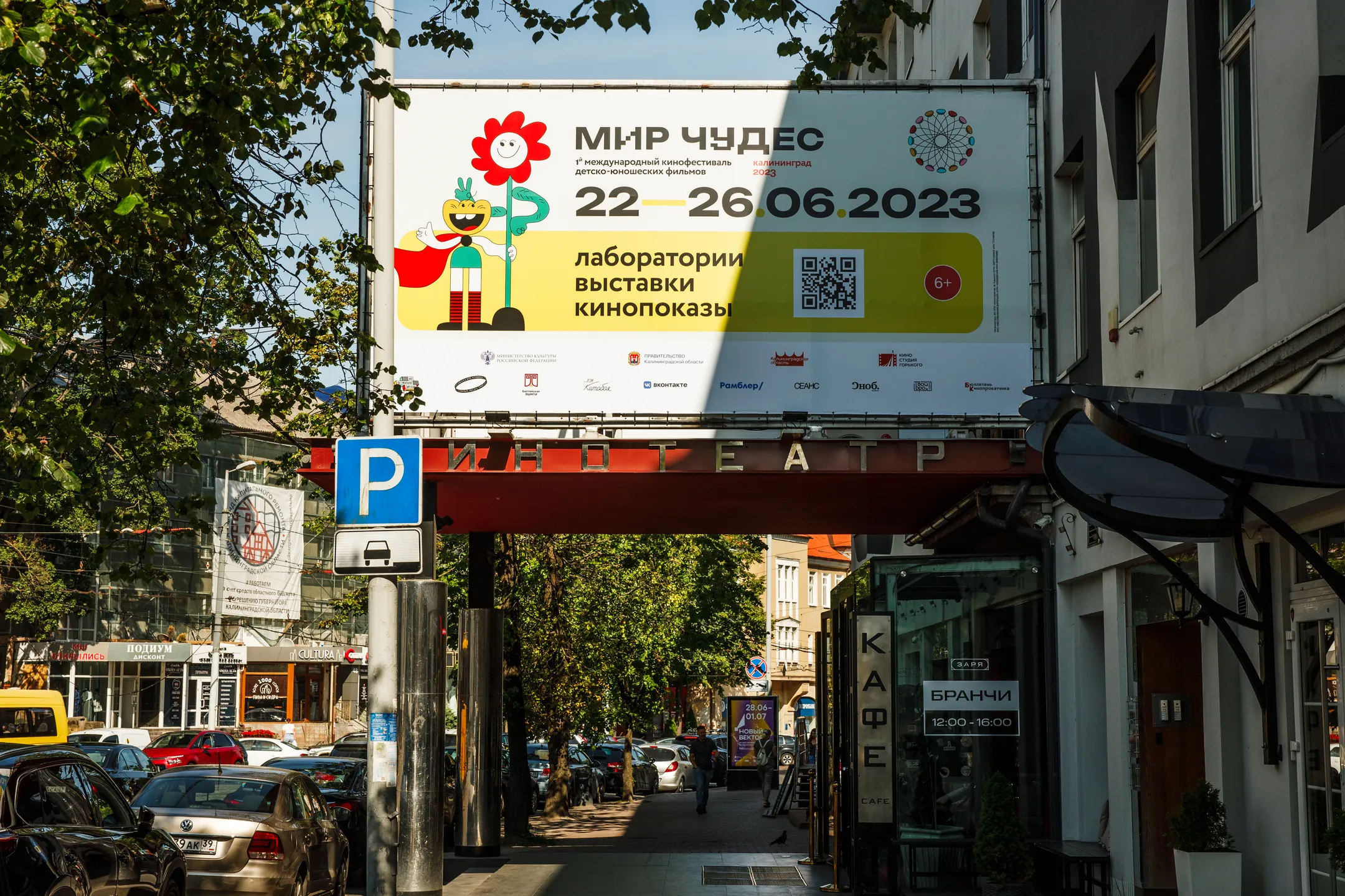 A festival banner strung across a street between buildings, above traffic and a theatre sign.