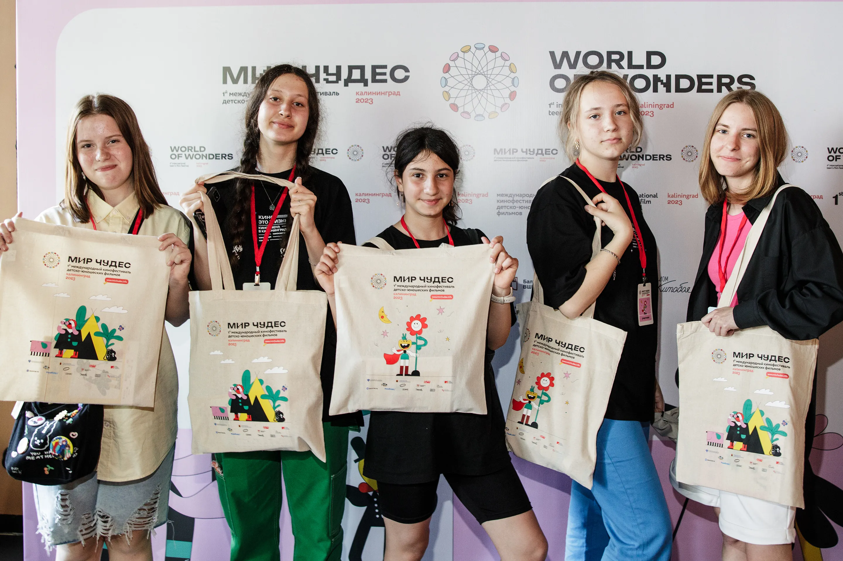 Five teenage festival attendees standing in front of the World of Wonders press wall, each holding a printed festival tote bag.