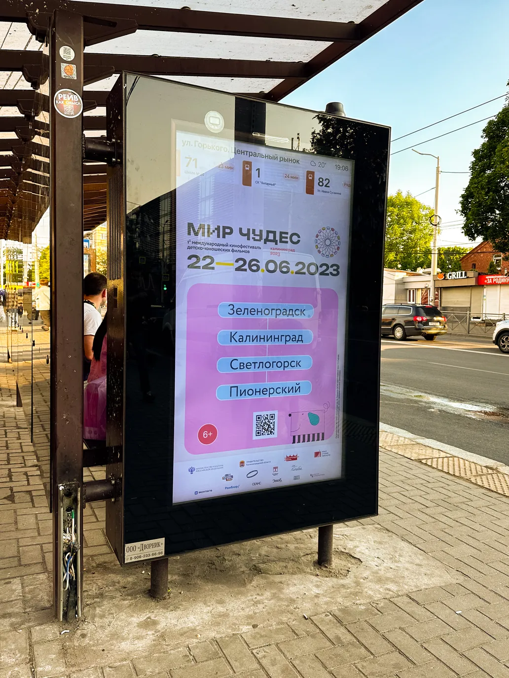A backlit citylight poster at a bus shelter, listing the four towns hosting the festival.
