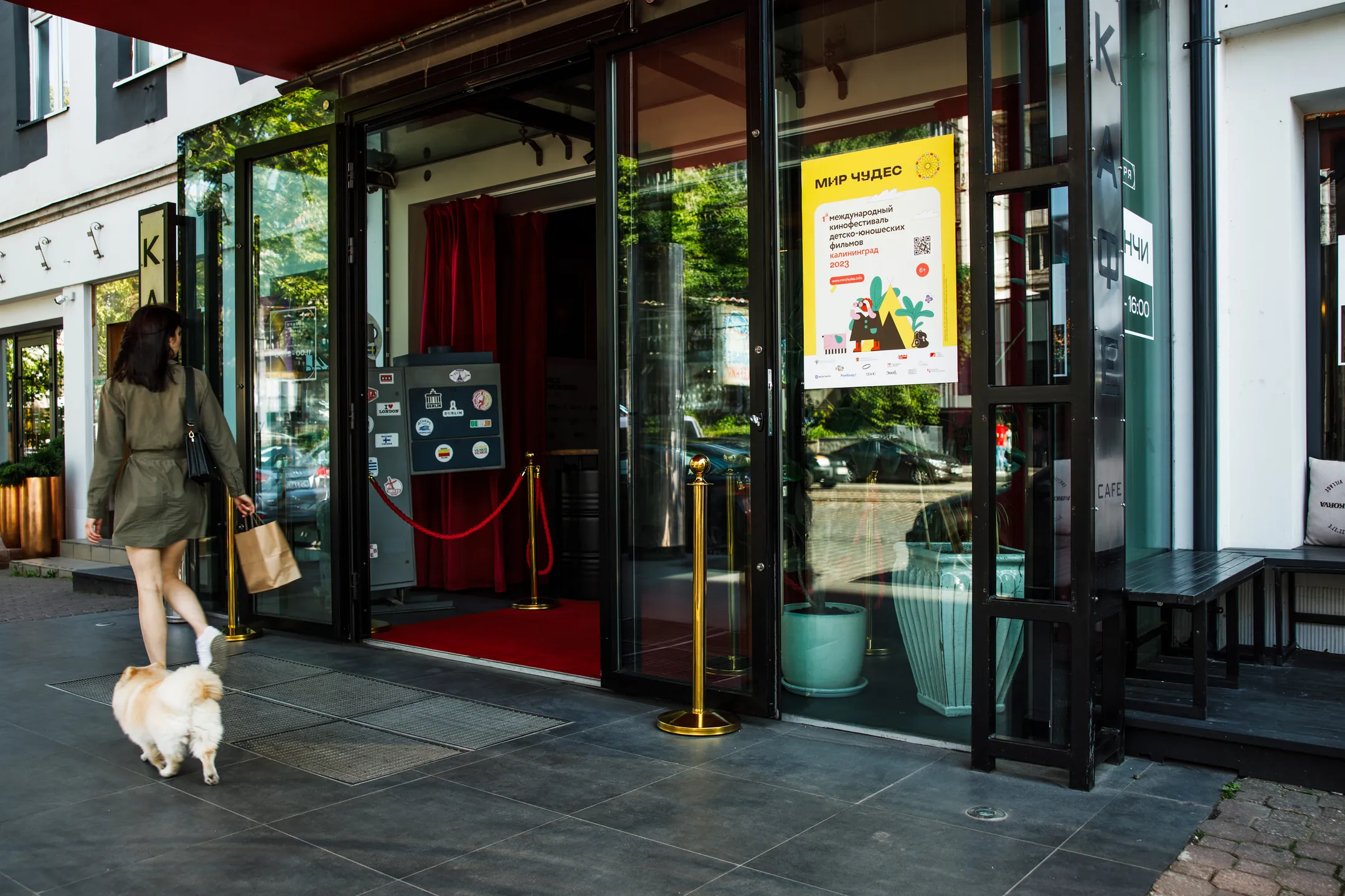 A woman walking a small dog past a shop doorway with the festival poster displayed inside the glass.